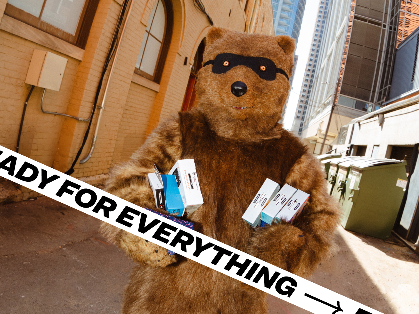 A bear mascot wearing a black mask, holding multiple product boxes in an alleyway, promoting readiness with the slogan 'Ready for Everything.